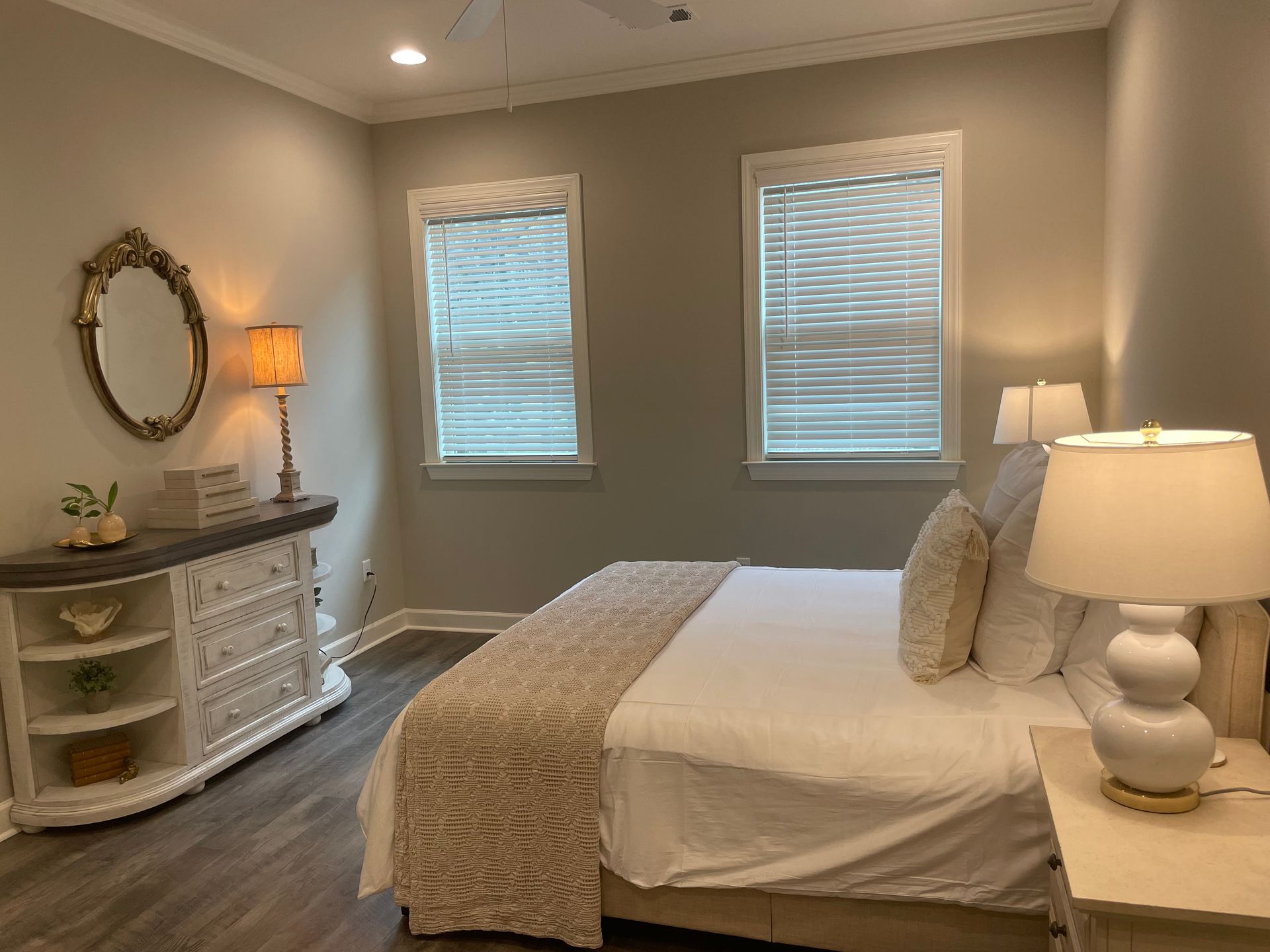 Bedroom with a white bed, dresser, and lamps. Two windows with blinds. Gray walls, wooden floor.