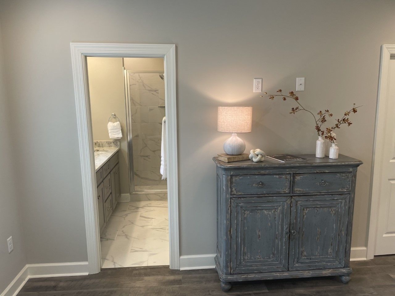 Bedroom with gray walls, cabinet, lamp, and open door to a bathroom with a shower and vanity.