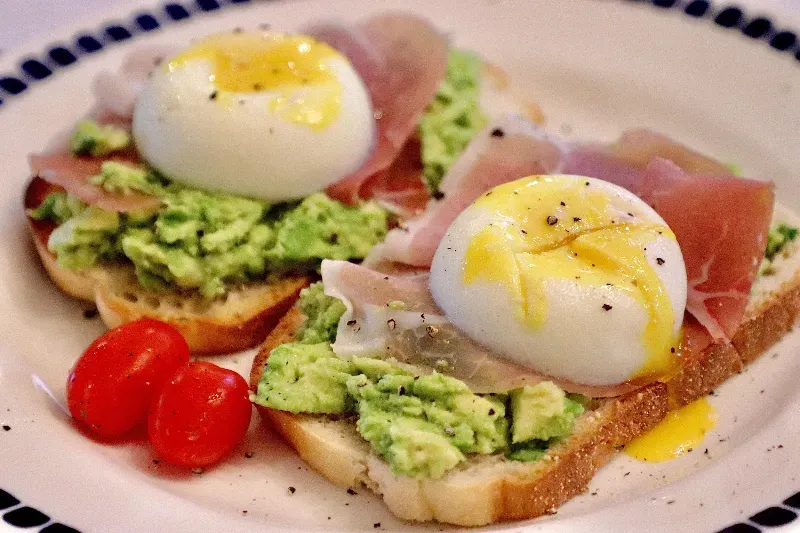 A close up of a plate of food with eggs , avocado , ham and tomatoes.