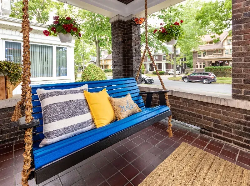 A blue porch swing with yellow and white pillows on a brick porch.