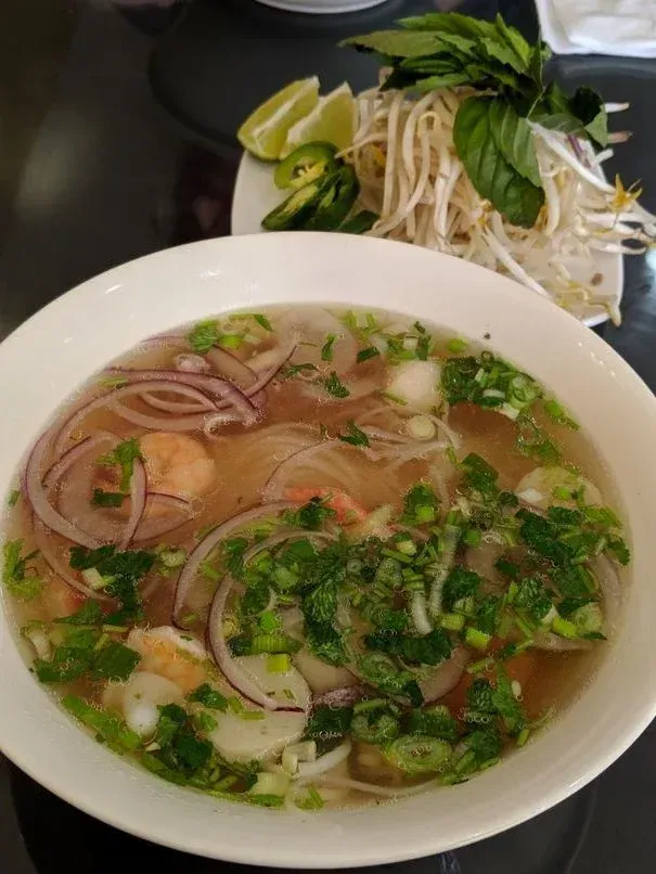 A bowl of soup with shrimp and onions on a table next to a plate of noodles.