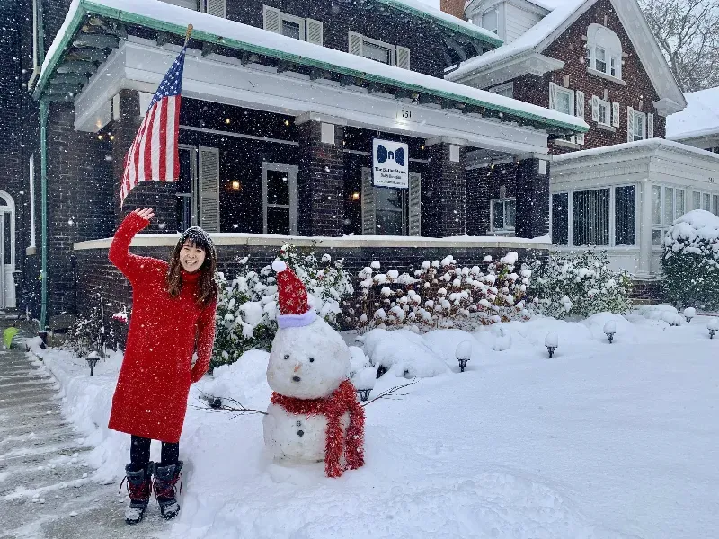 A woman in a red coat is standing next to a snowman in front of a house.