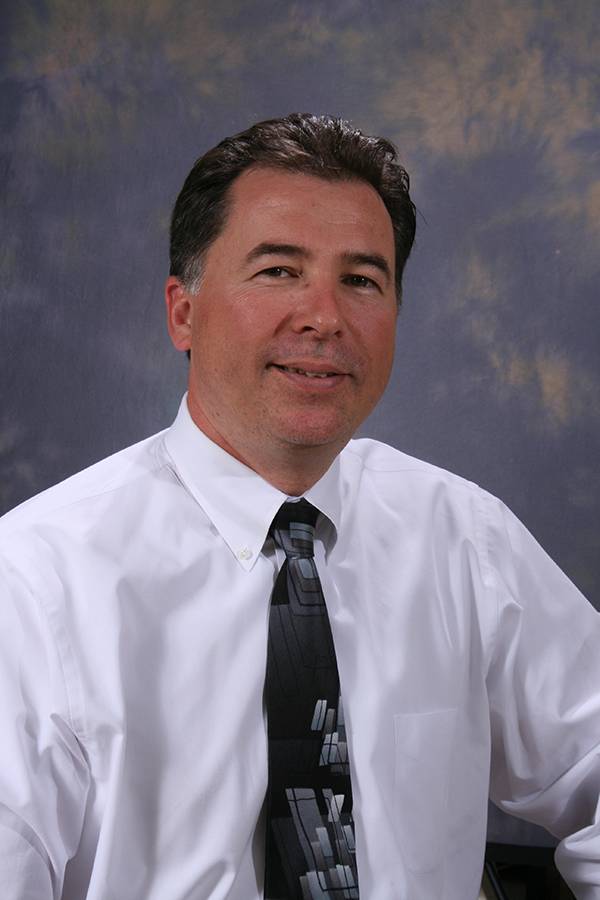 Man in a white shirt and tie smiles at the camera. Dark hair, neutral background.
