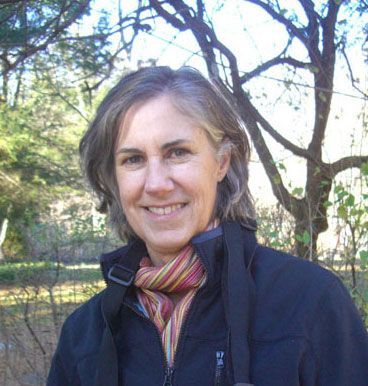 A smiling person with shoulder-length hair wearing a black jacket and a colorful striped scarf outdoors among trees.