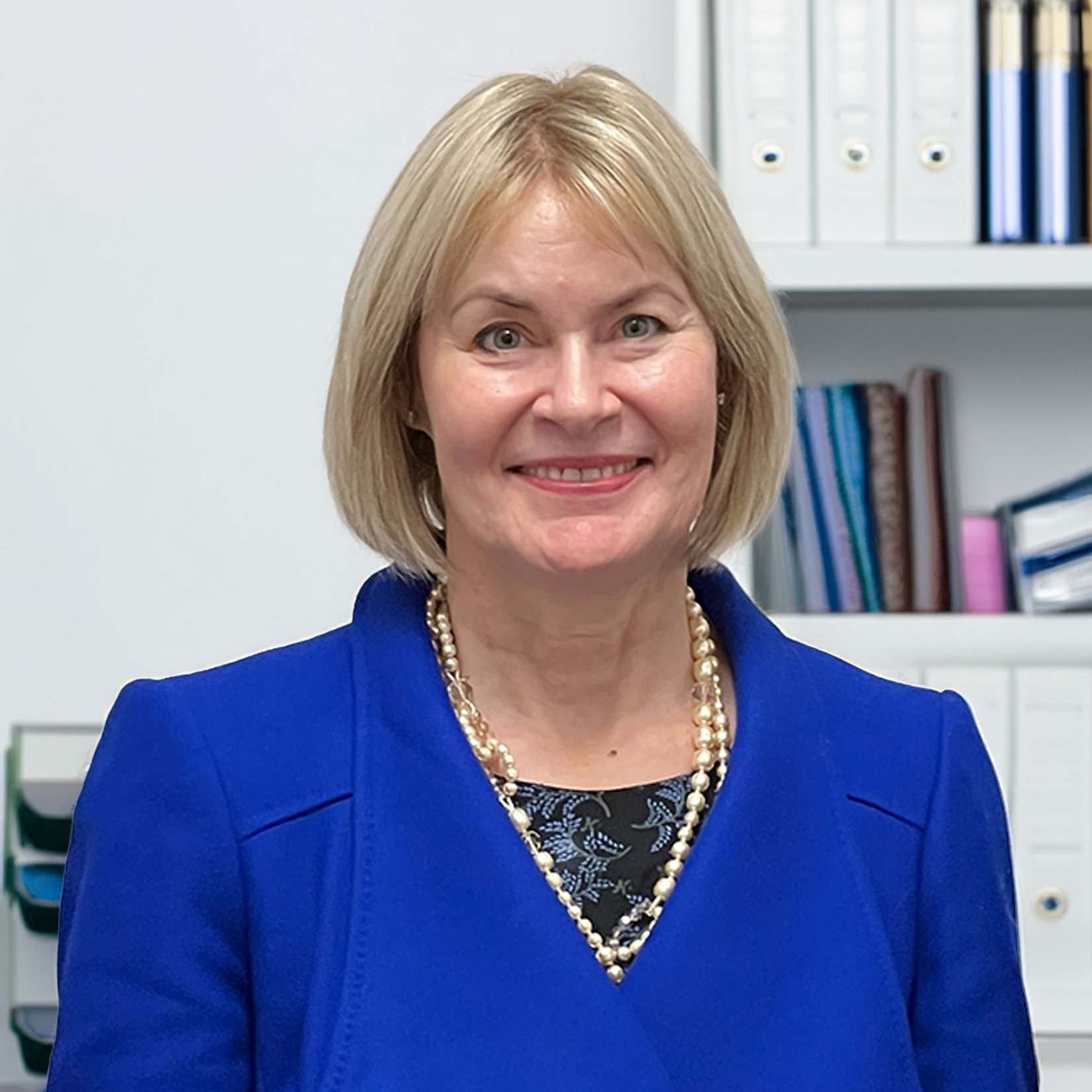 Woman with blonde bob smiles, wearing a blue jacket and pearl necklace, in an office.