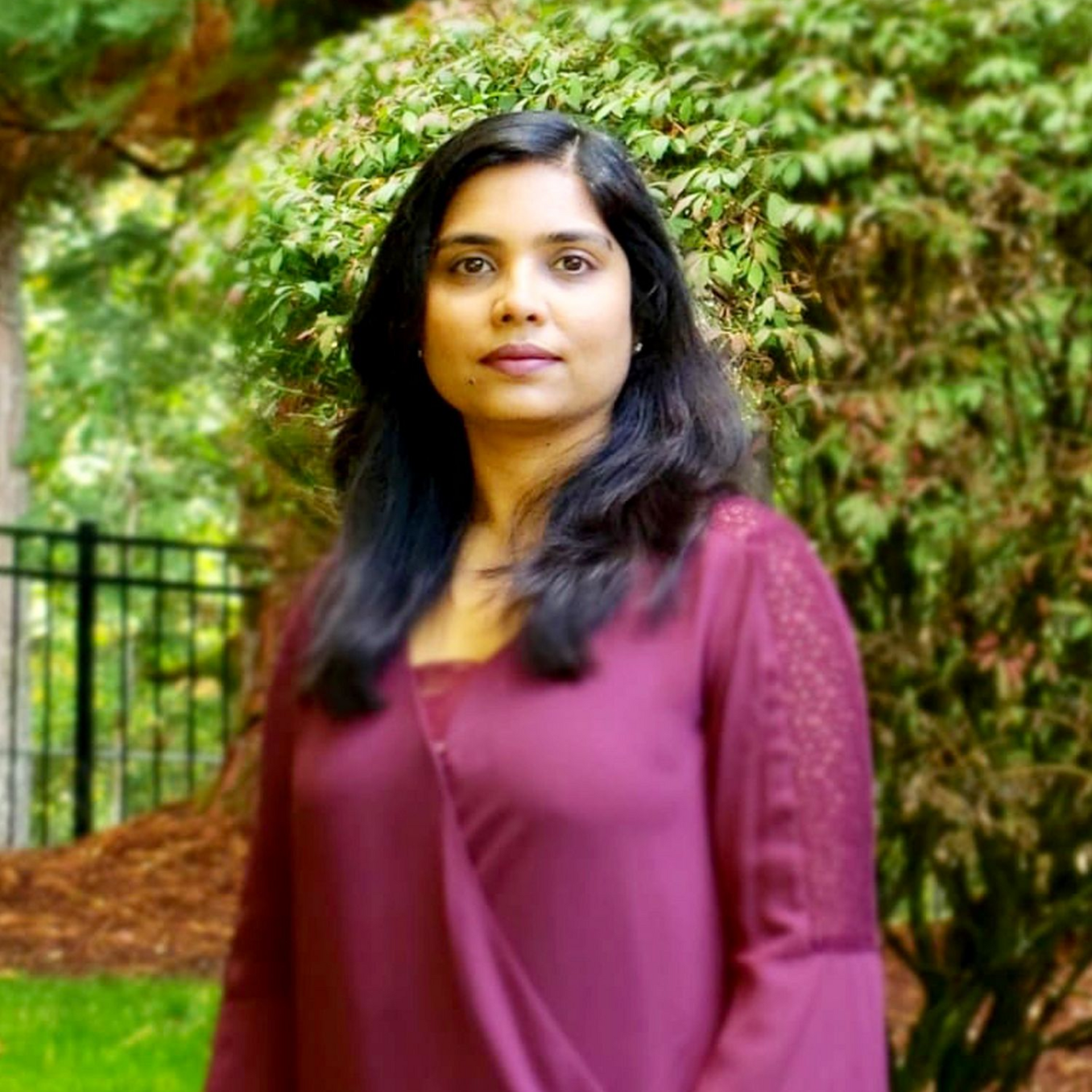 Woman in burgundy top, outdoors with greenery in the background.