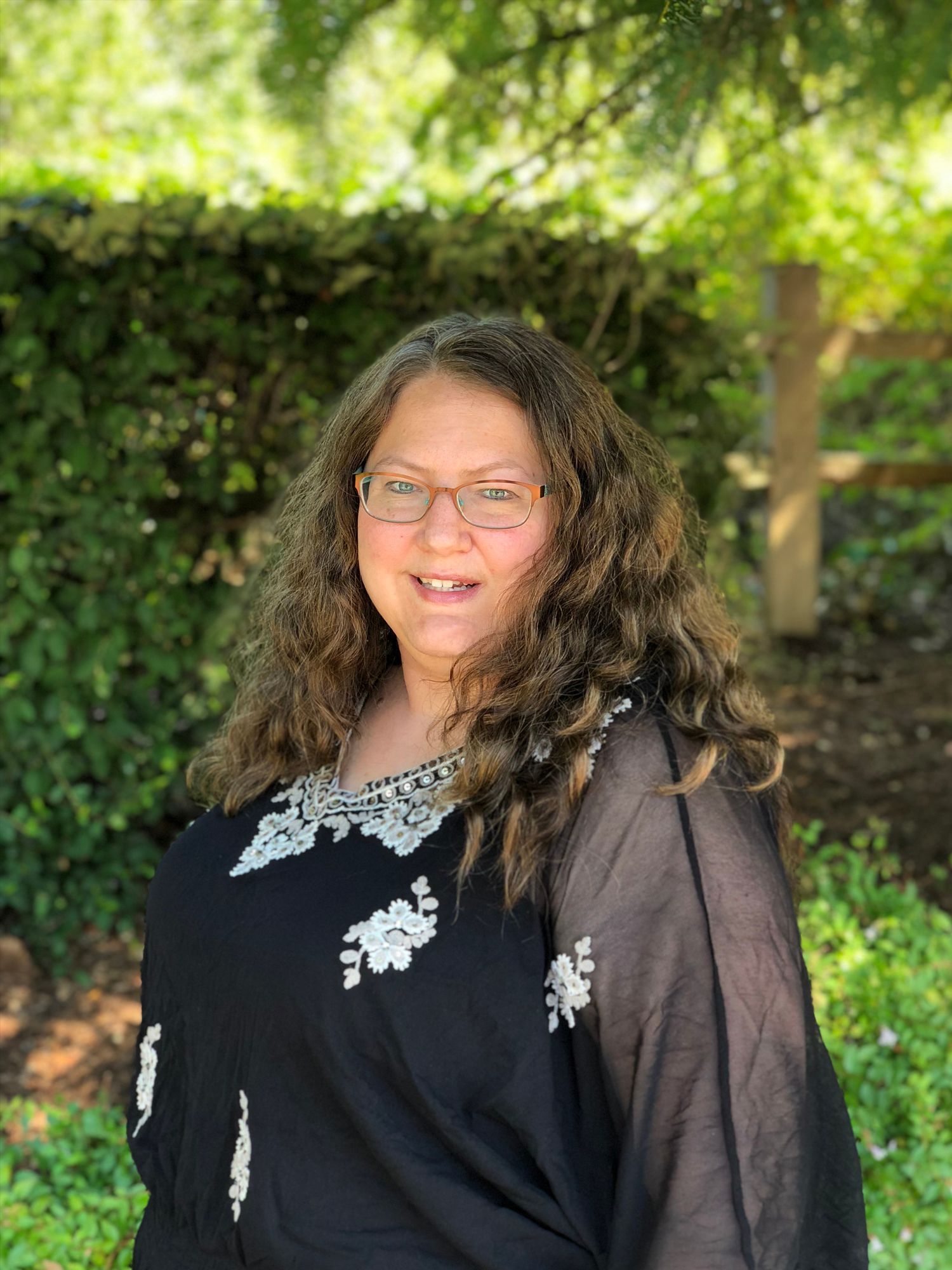 Woman with long, wavy brown hair and glasses, wearing a black top with white embroidery, smiling outdoors.