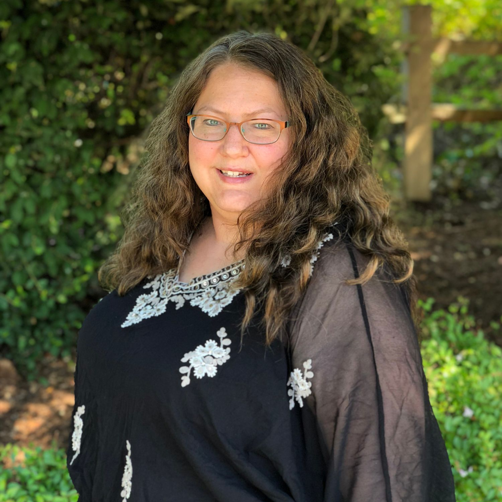 Woman wearing glasses and a black top with white embroidery smiles outdoors.