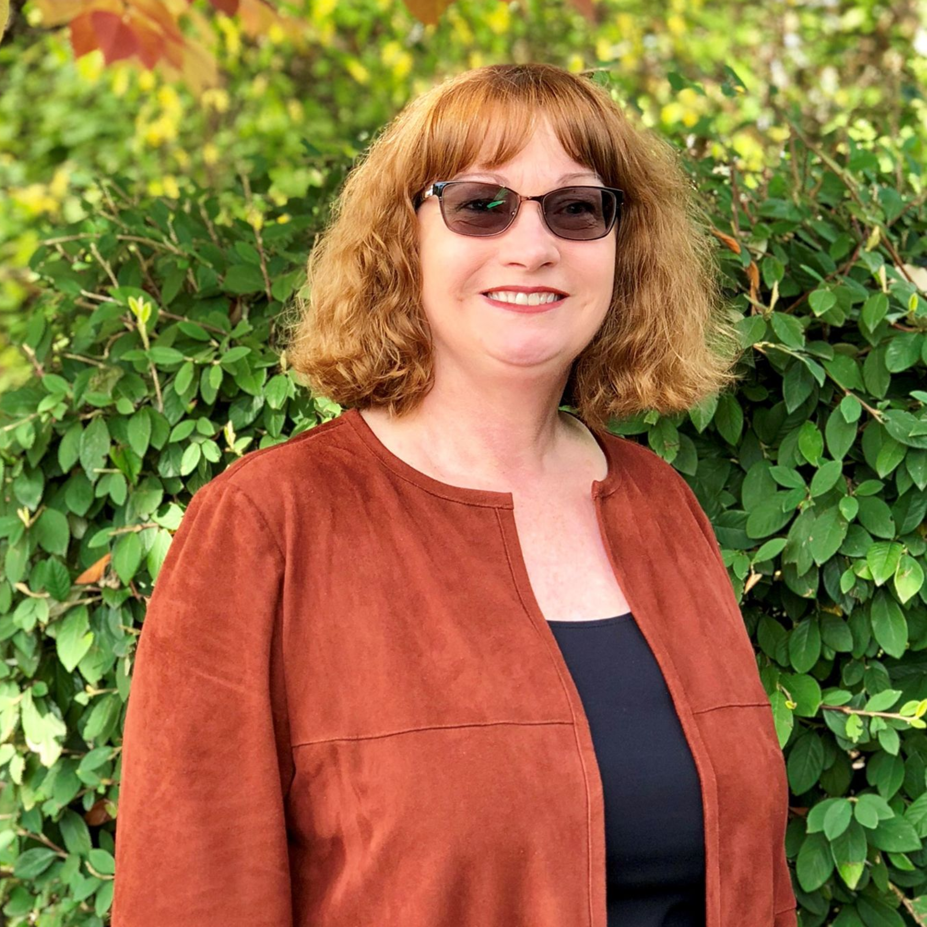 Woman with reddish hair and glasses smiles, wearing a rust-colored jacket over a black top, outdoors by green foliage.