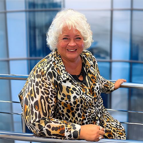 Woman in animal print shirt smiles, leaning on railing outside a glass building.