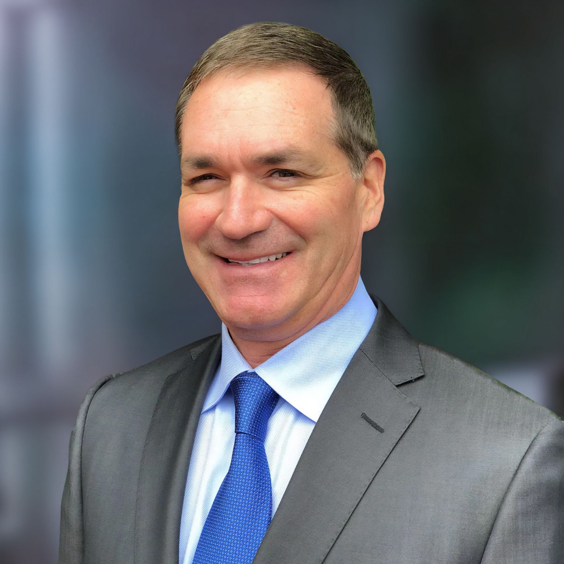 Man in gray suit with blue tie smiles in front of a blurred background.