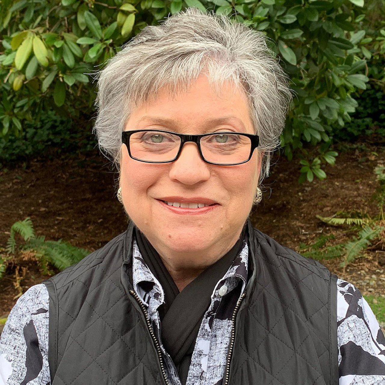 Woman with short gray hair and glasses smiles outdoors, wearing a black vest and patterned shirt.