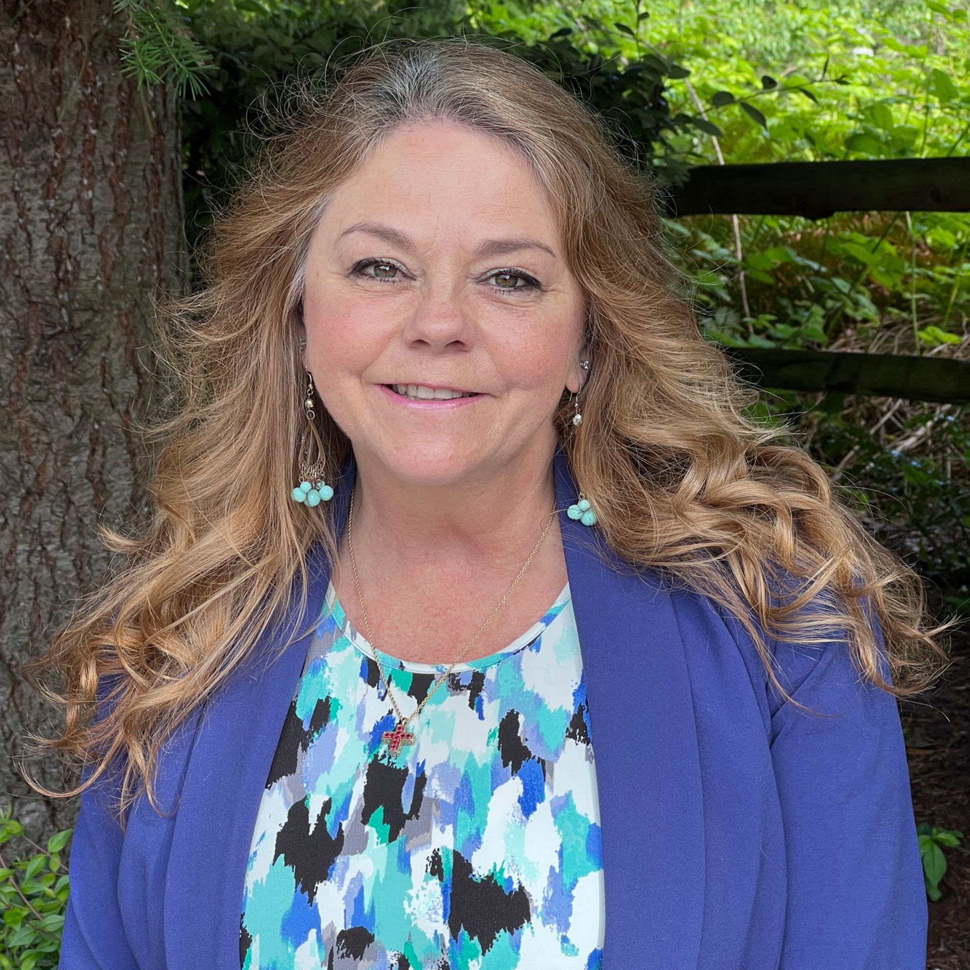 Woman with long wavy hair smiles, wearing blue jacket and patterned shirt, outdoors near a tree and fence.