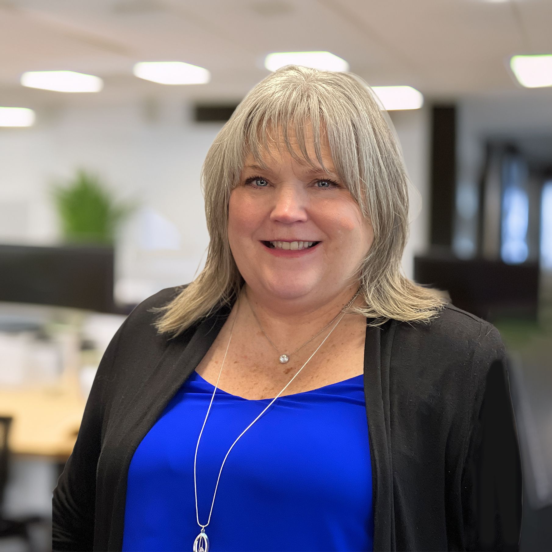 Woman with gray hair smiles, wearing blue top and black cardigan in office setting.