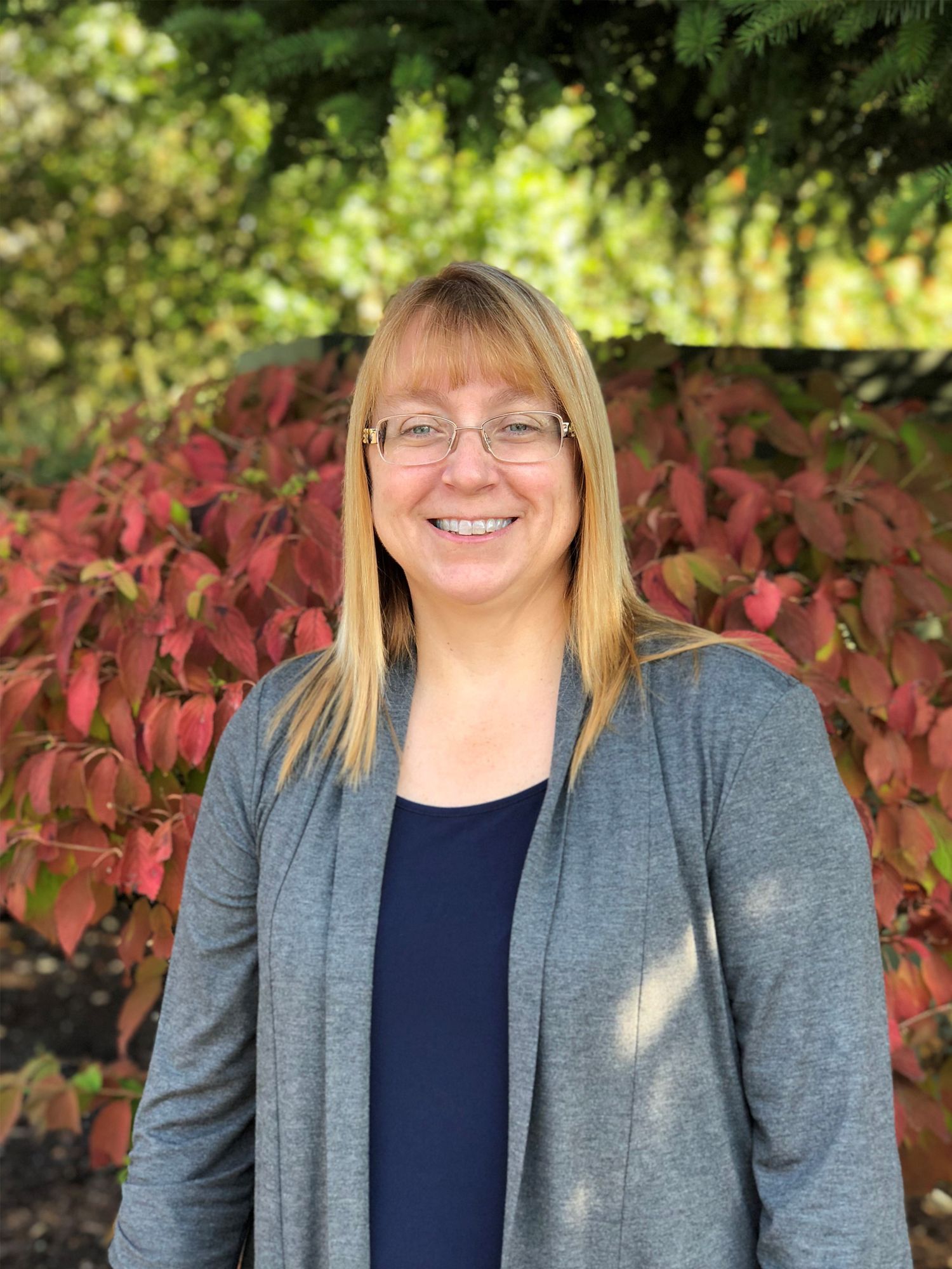 Woman with glasses smiling, wearing a blue shirt and grey cardigan, standing in front of red and green foliage.