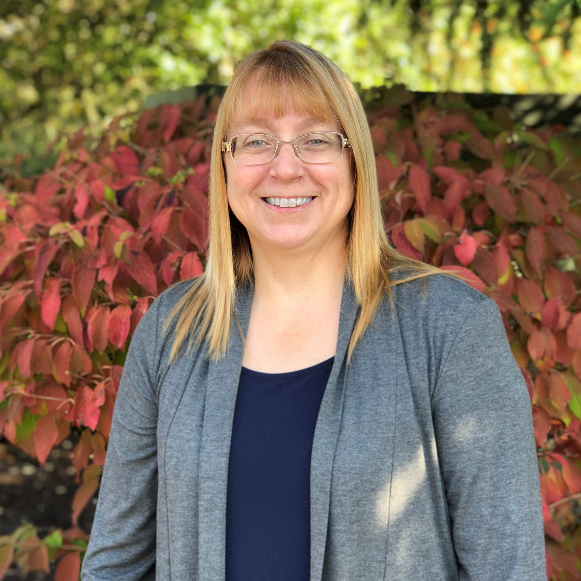 Woman with glasses smiles, wearing a gray cardigan and navy top, in front of red and green foliage.