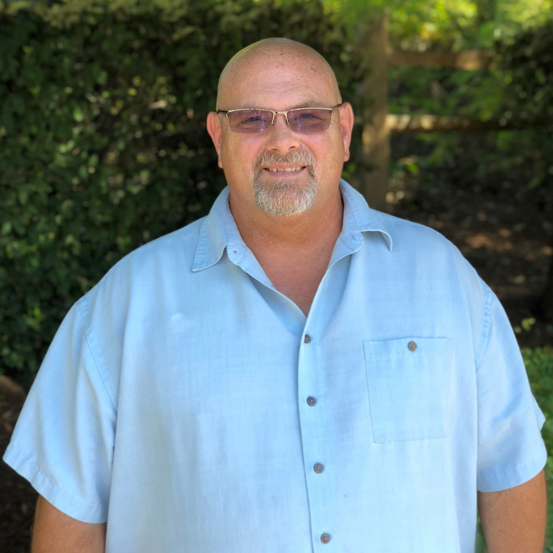 Man with glasses and goatee wearing a light blue shirt, smiling outdoors.
