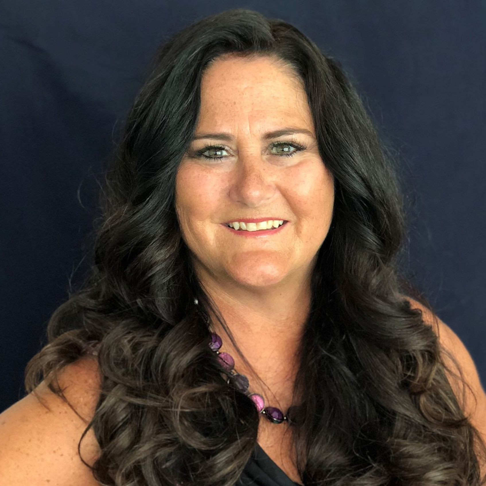 Woman with long, wavy dark hair smiles at the camera, wearing a black top and purple necklace.