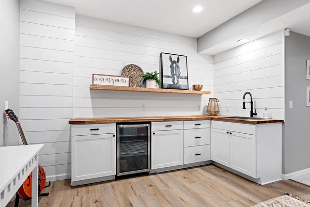 L-shaped home bar with white cabinets, wooden countertop, and shiplap walls; includes wine fridge, sink, and decor.