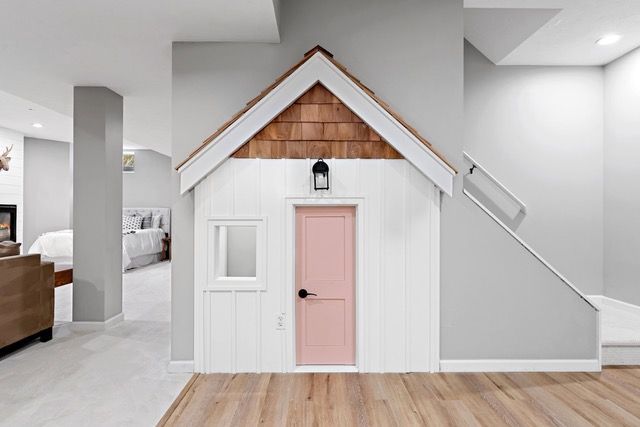 Playhouse built under stairs, white walls, pink door, wooden floor, brown roof shingles.
