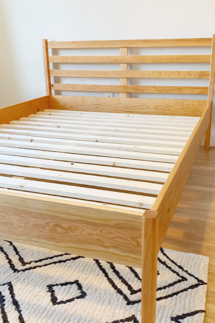 Wooden bed frame with a slatted headboard and base, set on a patterned rug.