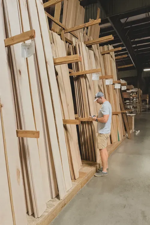 Man in a warehouse looking at wood planks. He’s taking notes, and wearing a hat and shorts.