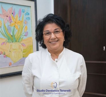 Una donna in camice bianco sorride in uno studio dentistico, con un dipinto floreale sulla parete alle sue spalle.