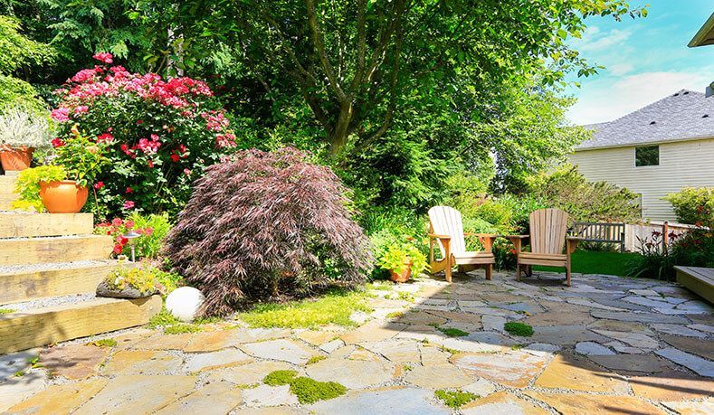 A backyard with a stone patio, chairs, flowers and a house in the background.