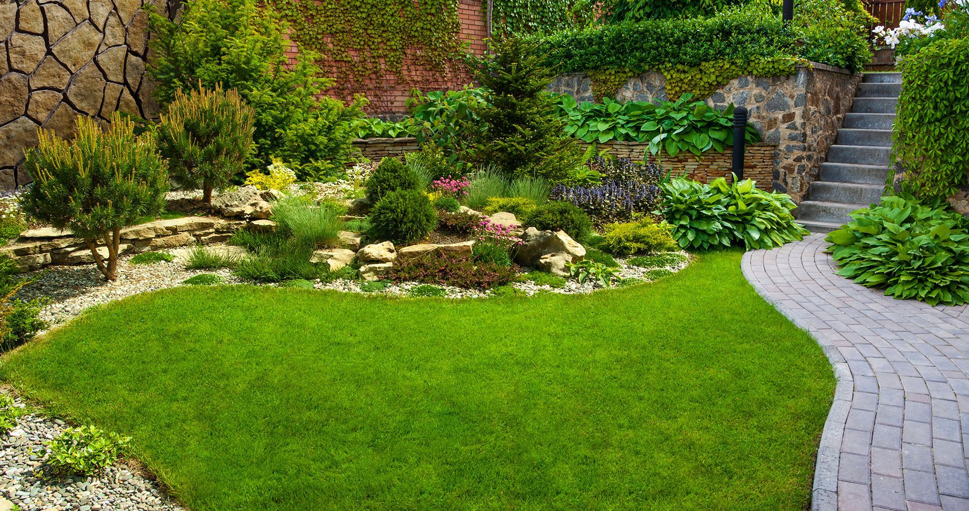 A lush green garden with a brick walkway leading to a stone wall and stairs.