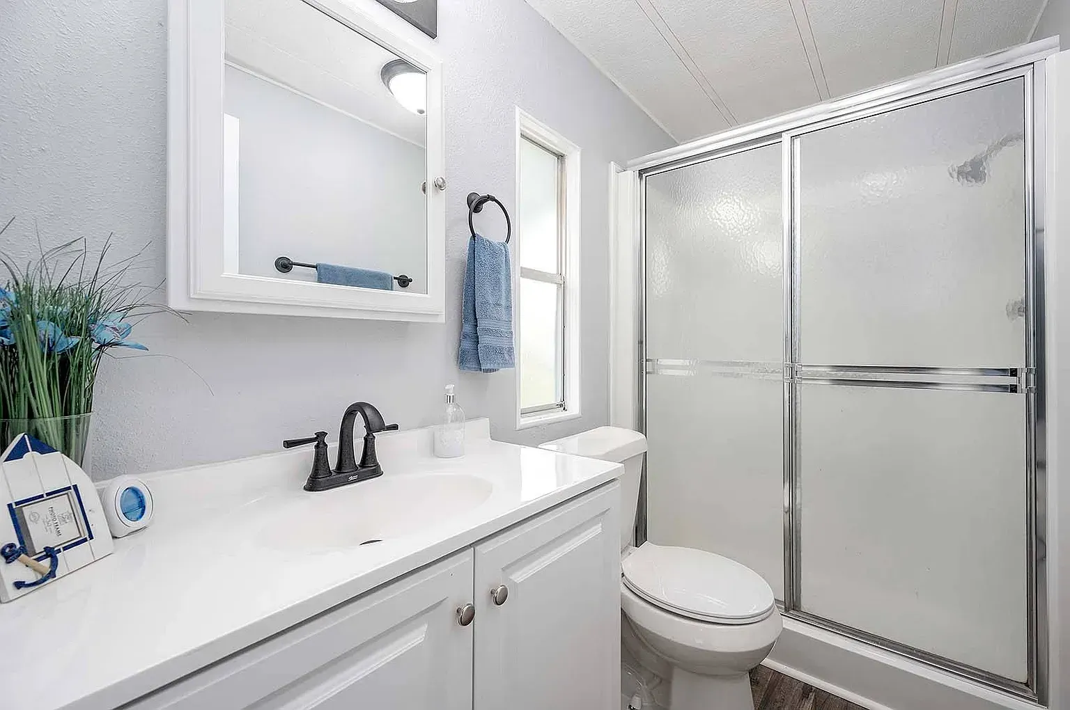 Bathroom with white vanity, toilet, shower, and window. Blue towel, flowers, and decor.