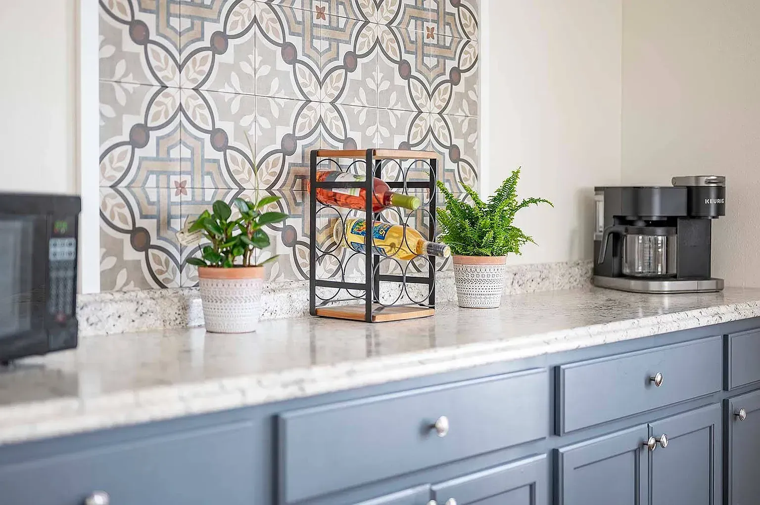 Kitchen countertop with patterned tile backsplash, wine rack, plants, microwave, and coffee maker. Blue cabinets.