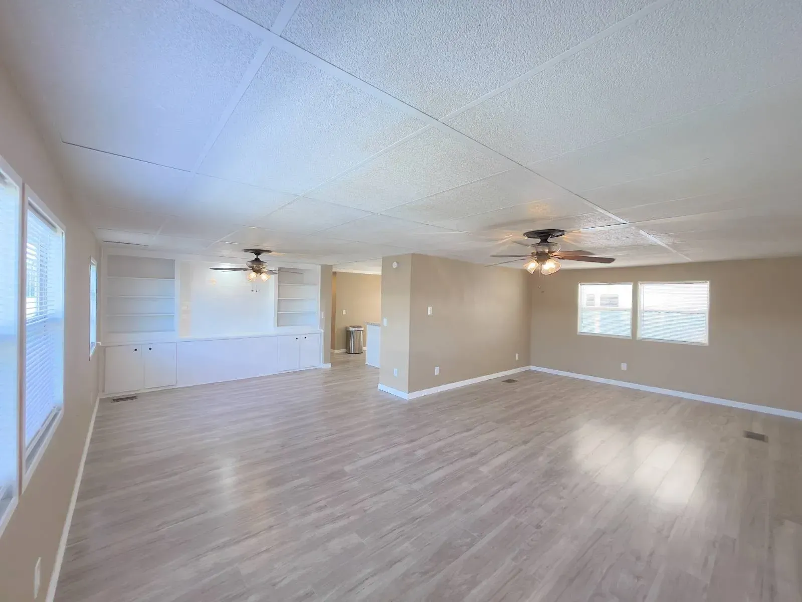 Spacious living area with wood flooring, two ceiling fans, windows, and beige walls.