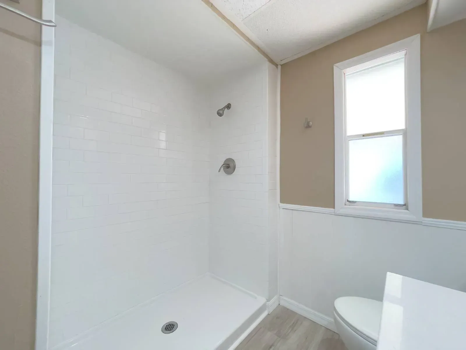 White-walled shower with showerhead, next to a toilet and window in a beige and white bathroom.