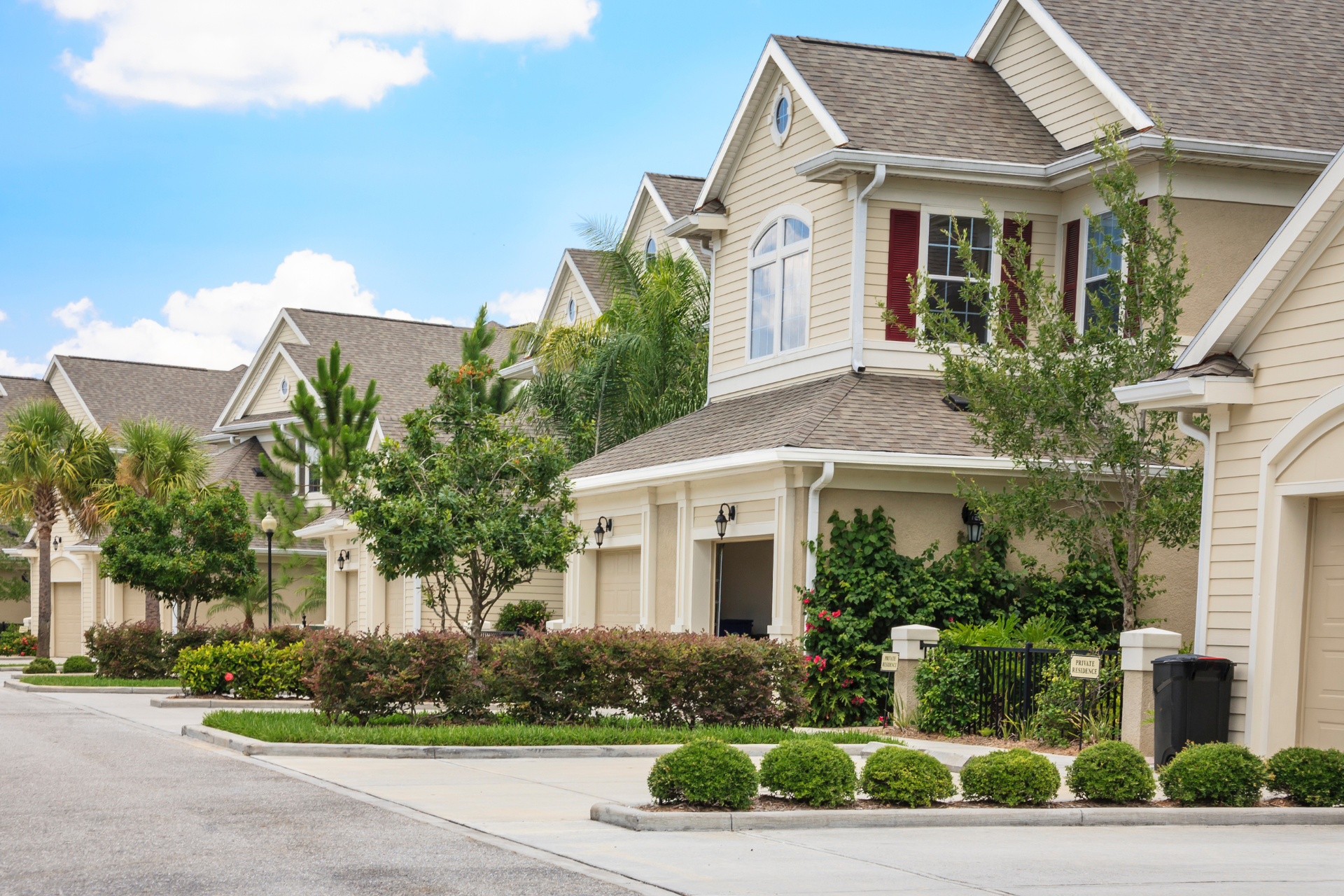 Suburban row of beige townhouses with manicured shrubs and a quiet driveway under a partly cloudy sky