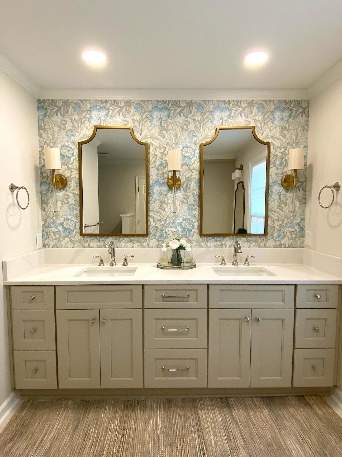Bathroom with double vanity, gold mirrors, floral wallpaper, and gray cabinets.