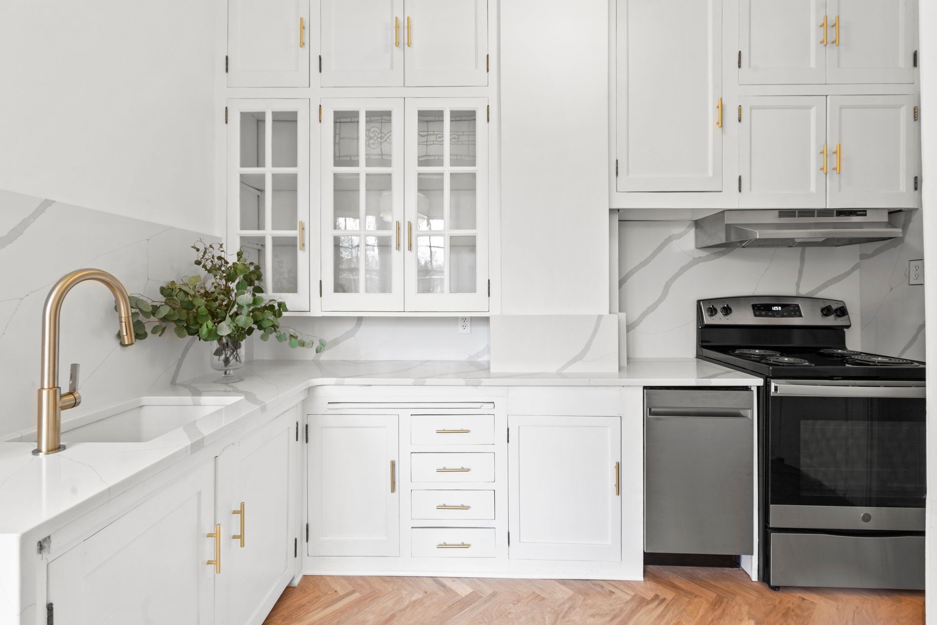 White kitchen with marble backsplash, stainless steel appliances, gold hardware, and wooden floor.