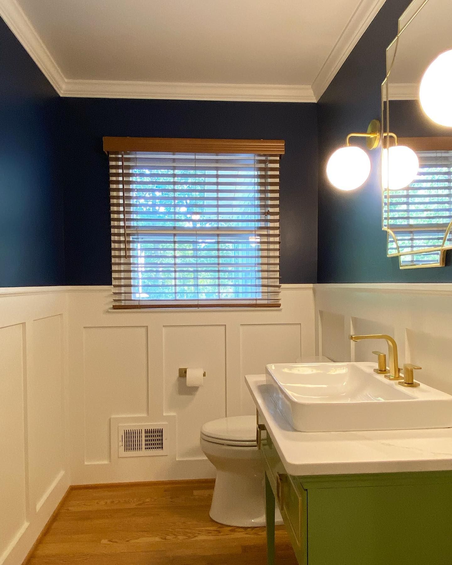 Bathroom with navy walls, white wainscoting, green vanity, and gold fixtures. Window with blinds.