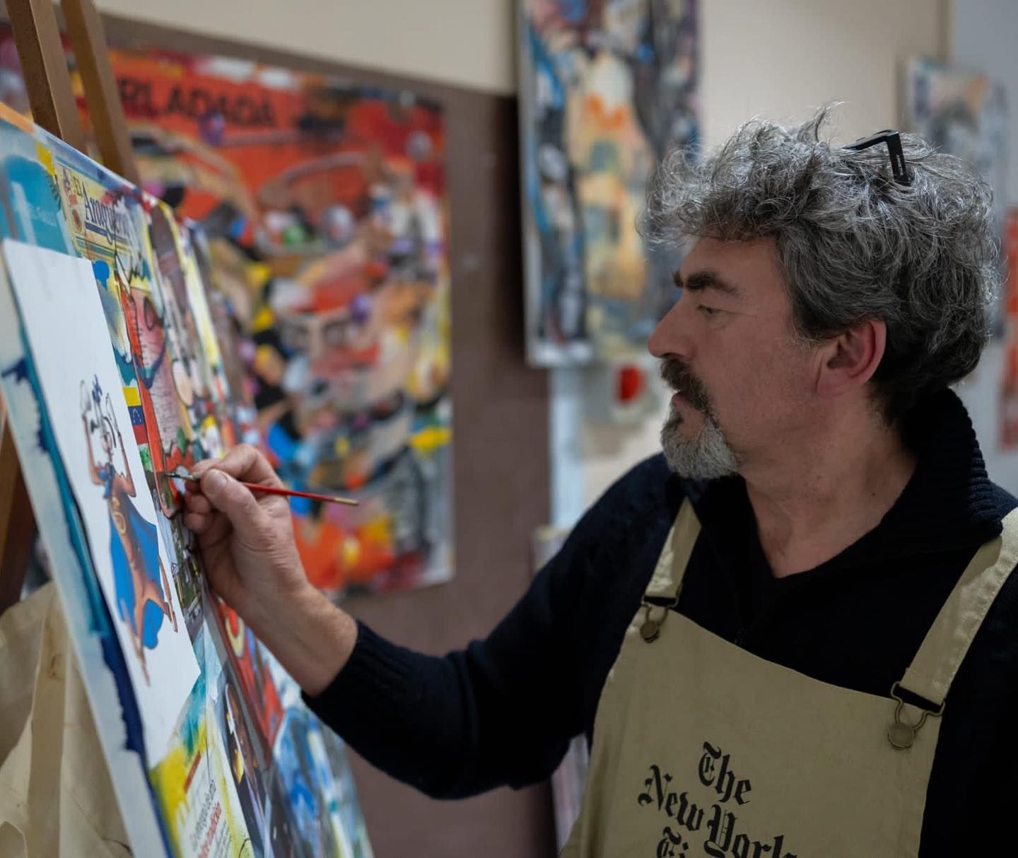 Un artista con indosso un grembiule color cachi dipinge su una tela in studio, circondato da altre opere d'arte colorate.
