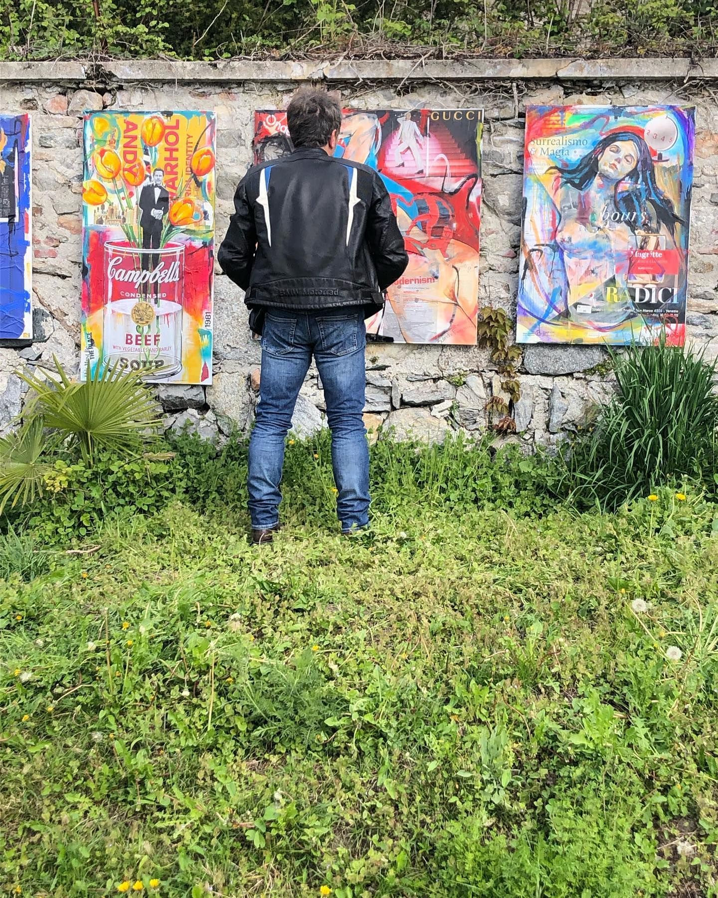 In un giardino, una persona con indosso una giacca di pelle nera e jeans è di fronte a un muro di pietra decorato con stampe artistiche colorate incorniciate.