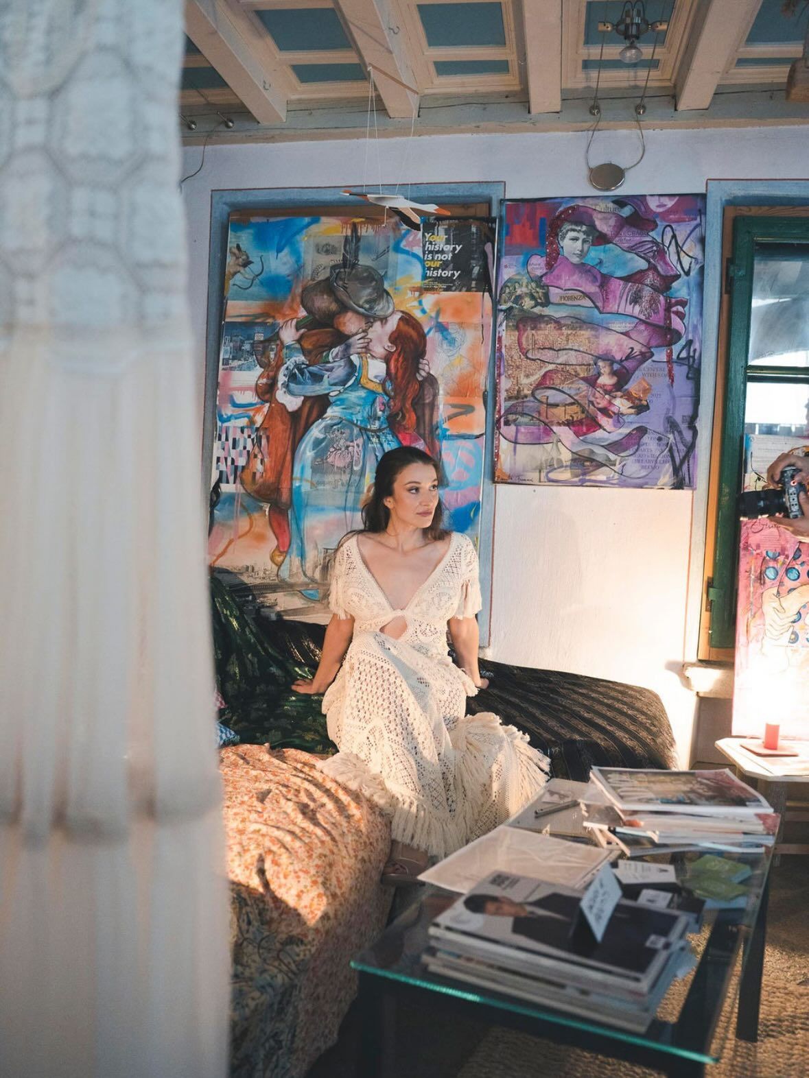 Una persona con indosso un abito all'uncinetto color crema è seduta su un divano di fronte a quadri colorati in una stanza con un soffitto rustico.