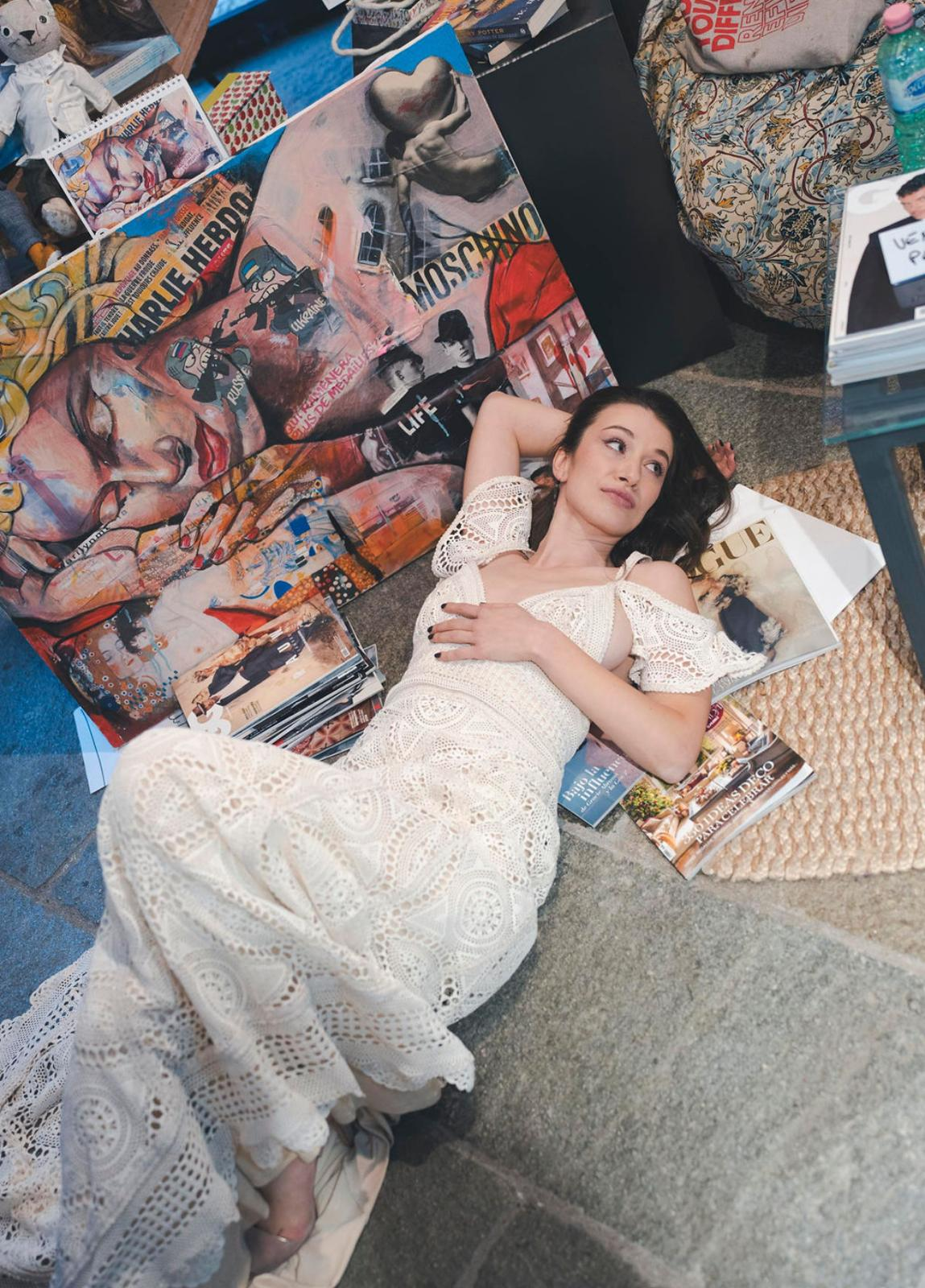 Una persona che indossa un elaborato abito di pizzo bianco lungo fino ai piedi si trova in uno studio creativo e disordinato, ricco di opere d'arte eclettiche.