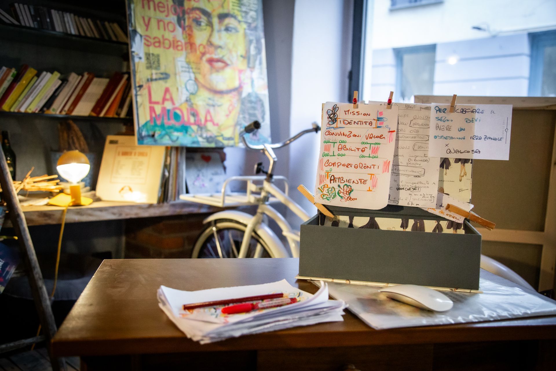 Una scrivania con penne su fogli di carta, una scatola con appunti scritti a mano e spillati sopra, una bicicletta e un poster di Frida Kahlo su una parete.