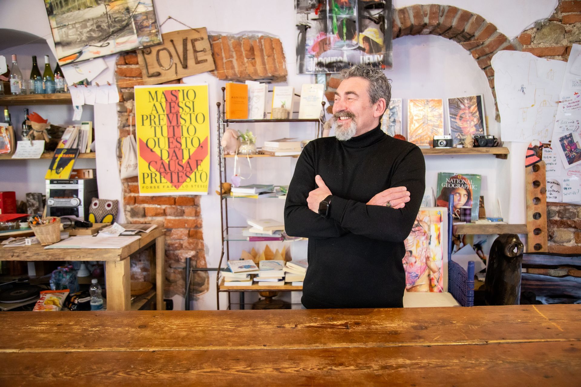In una libreria eclettica e ben illuminata, un uomo con la barba è in piedi con le braccia incrociate dietro un bancone di legno.