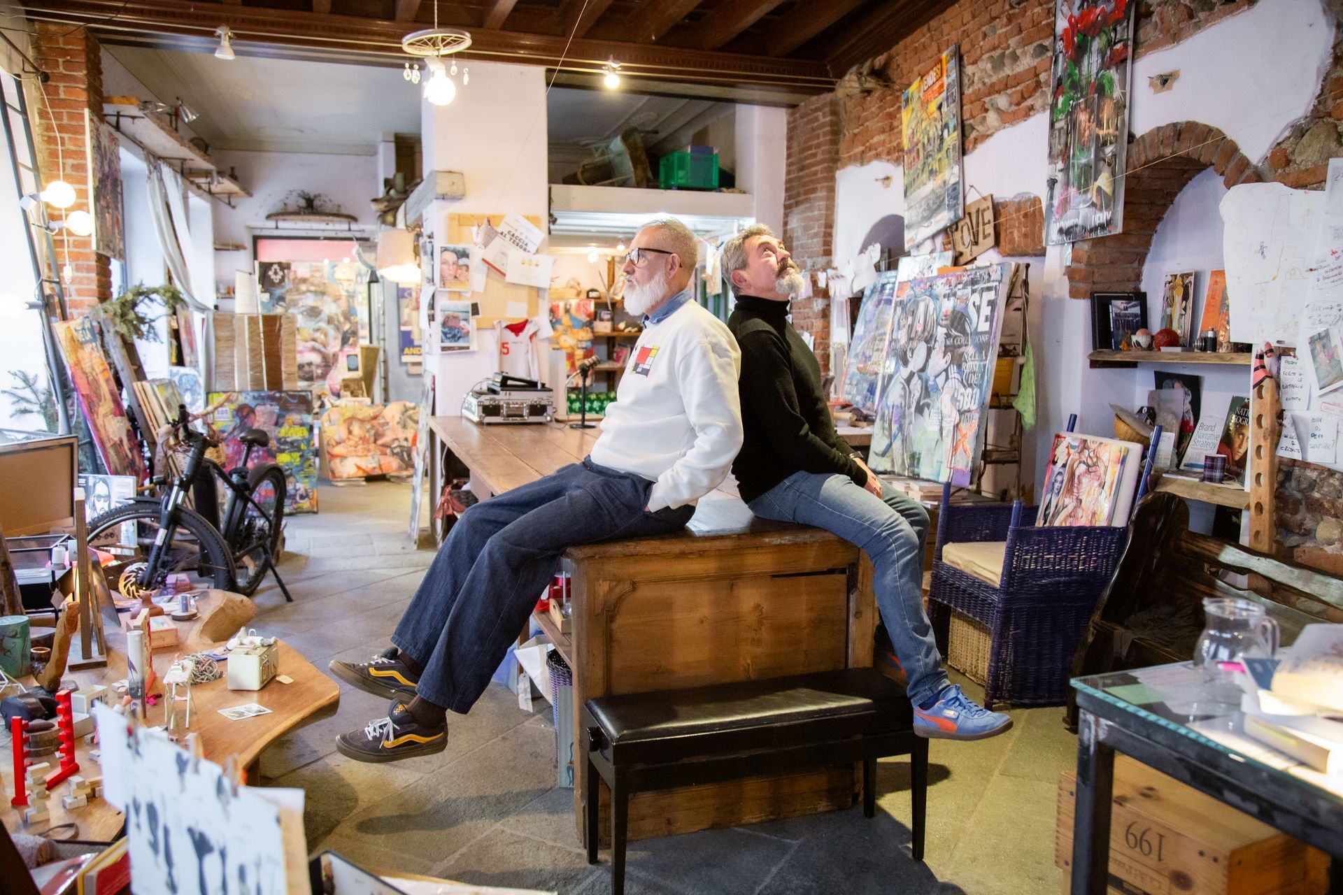 In uno studio ben illuminato e pieno di materiali artistici e poster, due persone siedono schiena contro schiena su un bancone di legno.