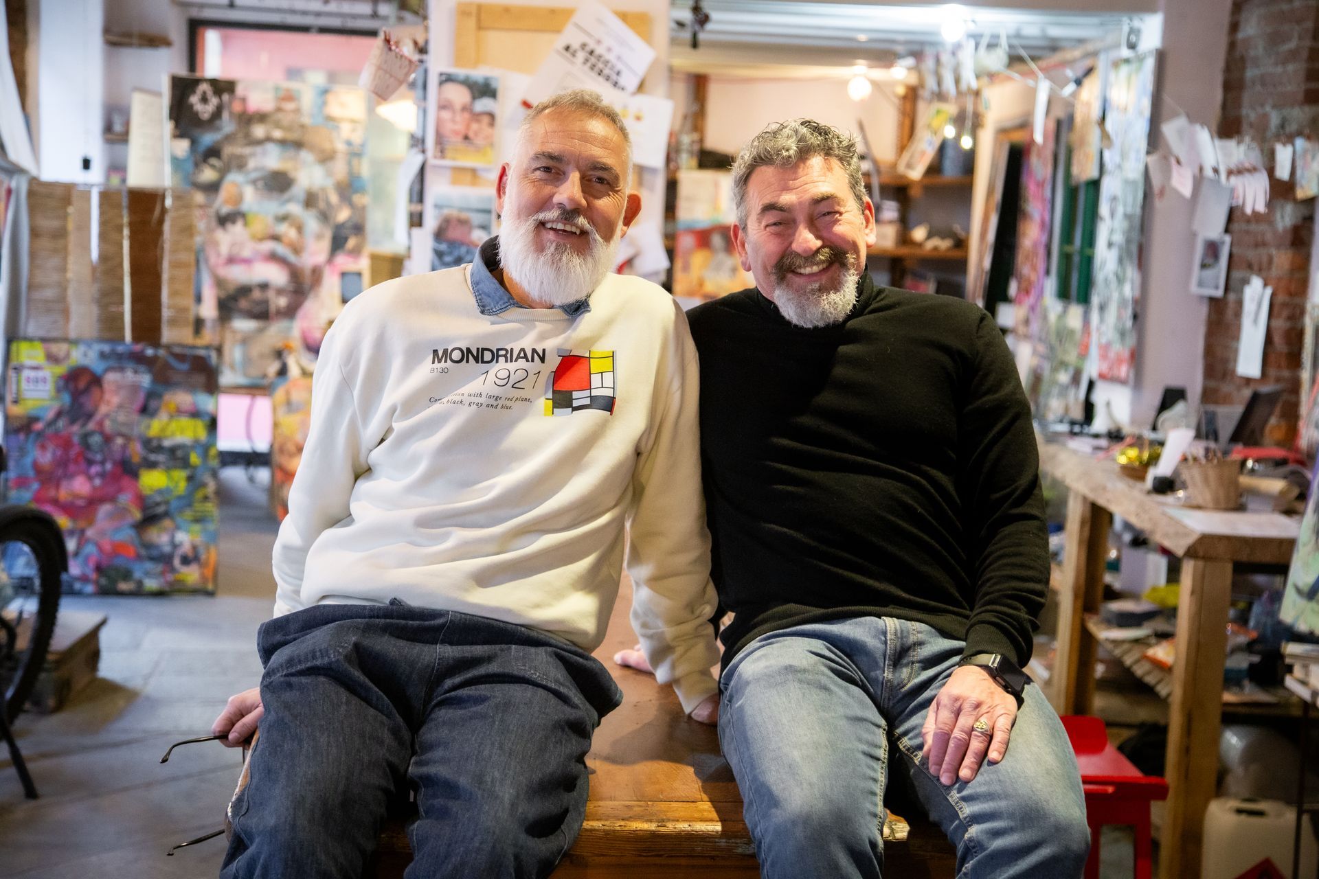 Due uomini barbuti dal sorriso amichevole siedono fianco a fianco in uno studio d'artista disordinato, pieno di quadri e materiali.