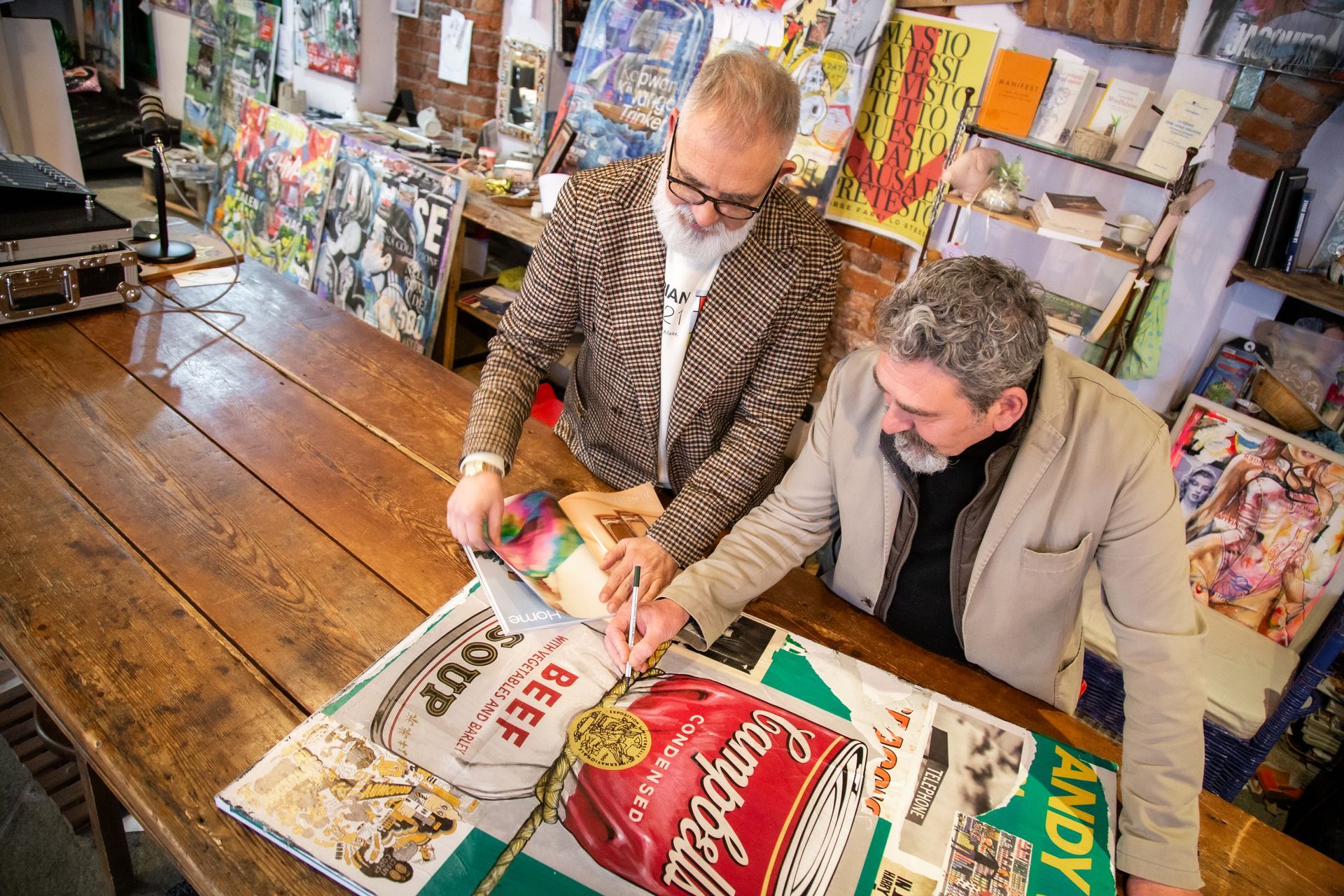 In uno studio d'arte, due persone lavorano insieme a un collage attorno a un tavolo di legno, raffigurante la grafica di una lattina di zuppa Campbell.