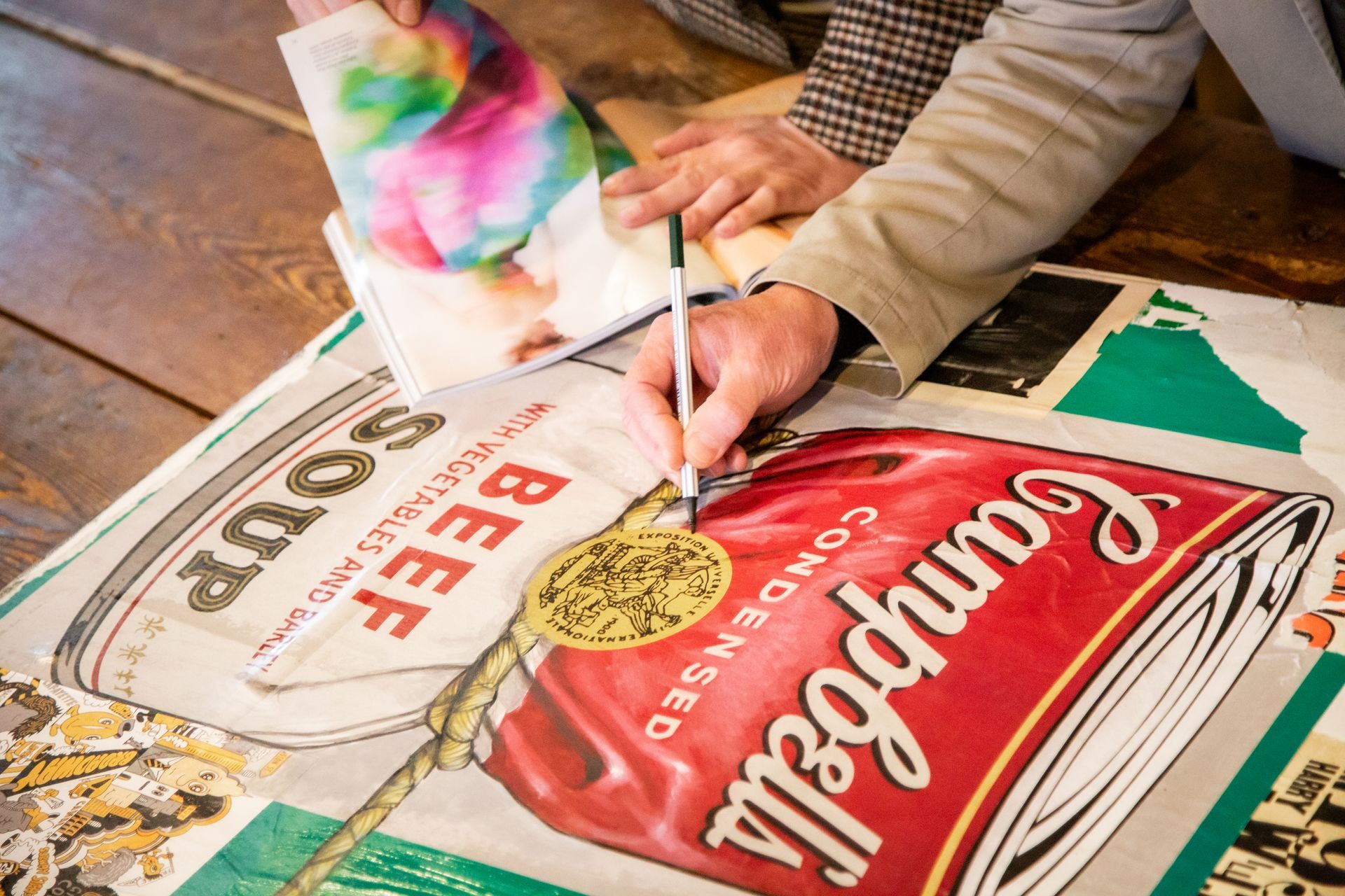 Una persona usa una penna per disegnare su una grande stampa raffigurante il disegno di una lattina di zuppa di manzo Campbell's, posizionata su un pavimento di legno.