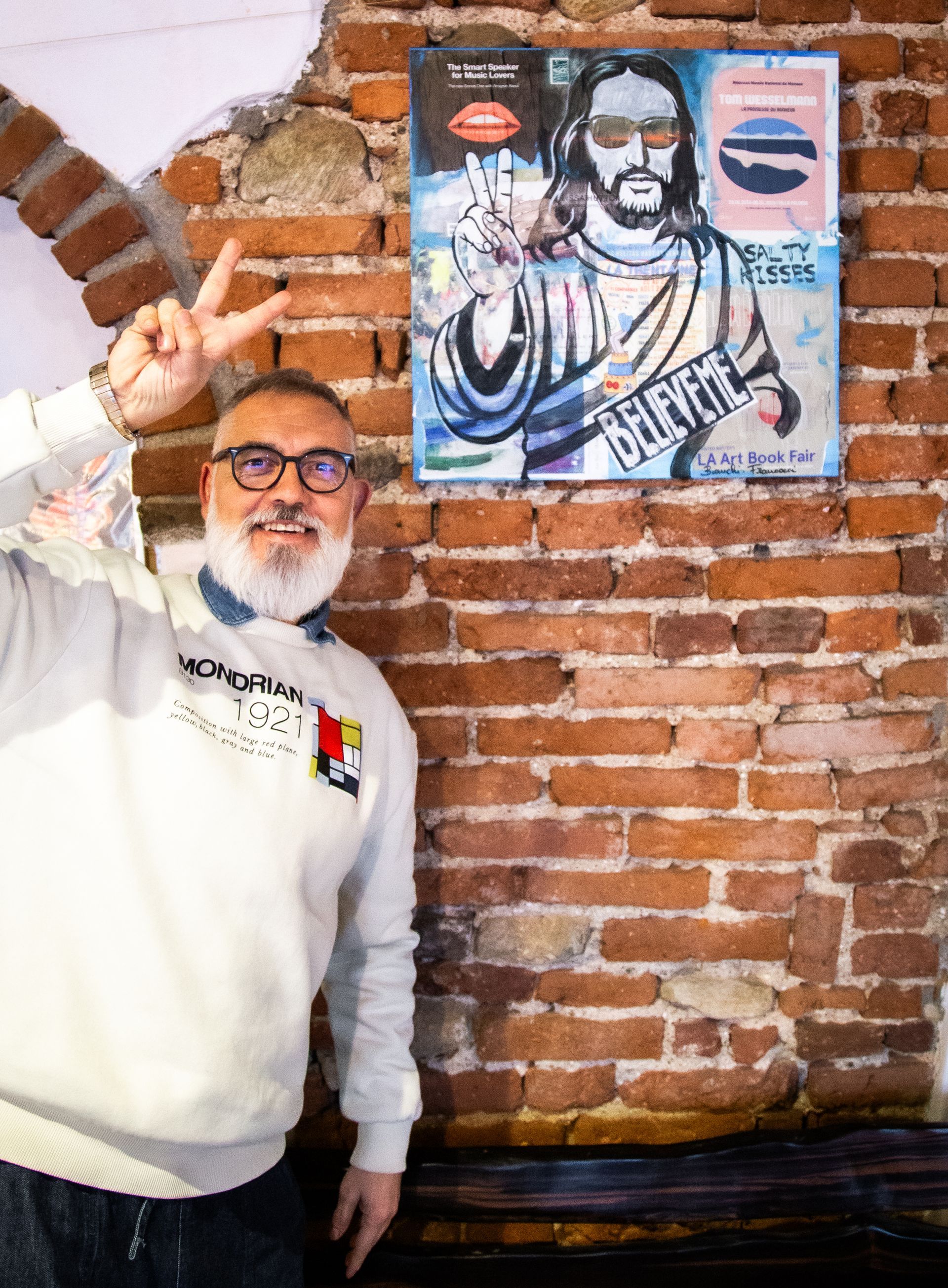 Un uomo con la barba bianca e gli occhiali posa, facendo il segno della pace accanto a un dipinto di Gesù su un muro di mattoni.