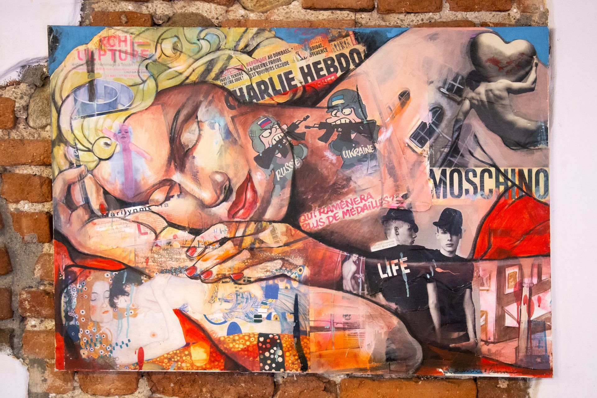 Collage a tecnica mista su muro di mattoni raffigurante una donna addormentata, riferimenti a Charlie Hebdo, il logo Moschino e figure armate.