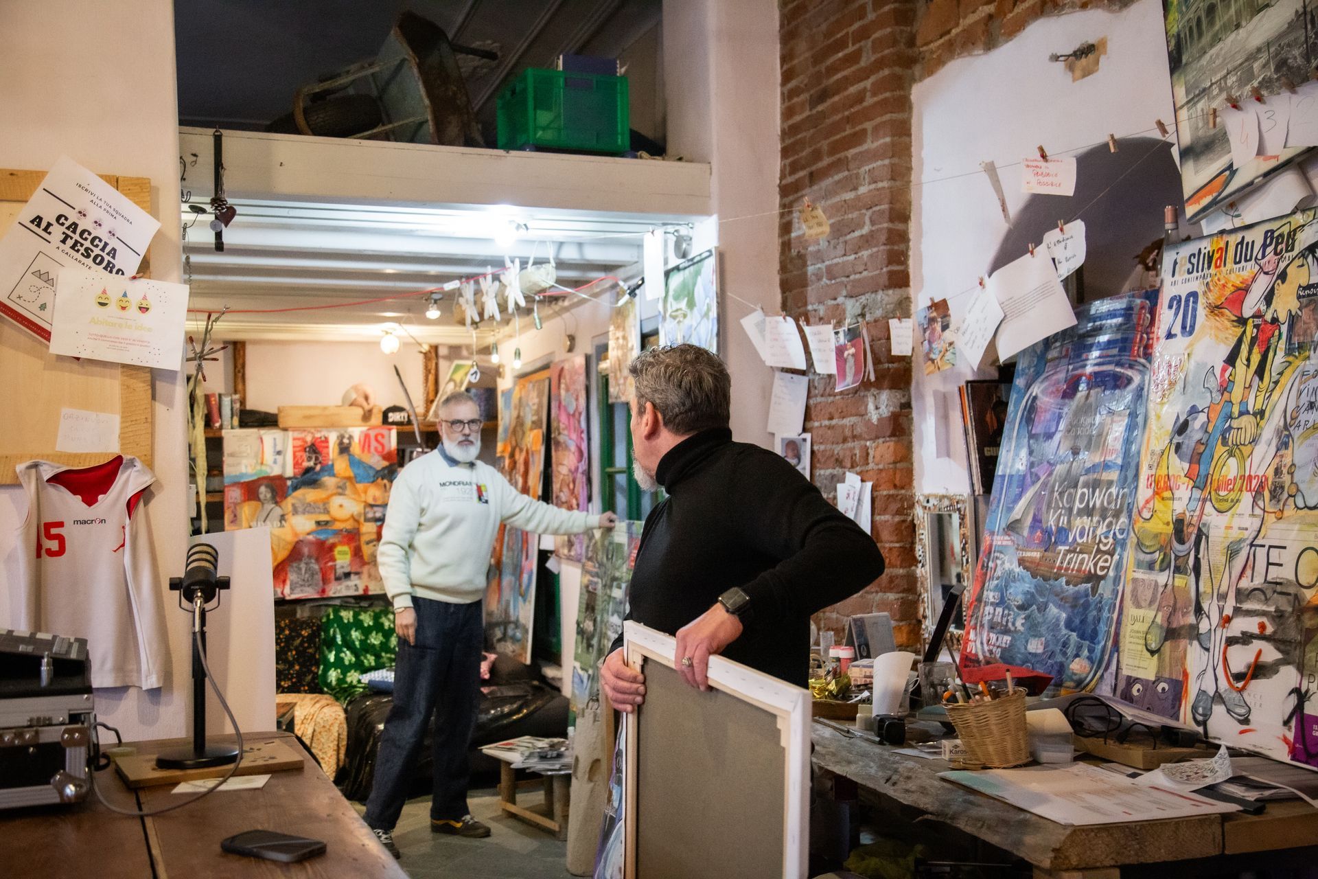 Due persone in uno studio d'arte, una in piedi vicino a un'opera d'arte e l'altra che tiene in mano una tela incorniciata vicino a un tavolo di legno.